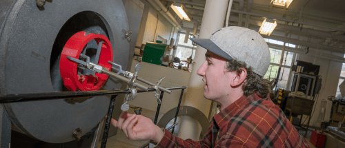 Student in a plaid shirt and gray cap adjusts a metal apparatus attached to a red component inside an industrial furnace in a Norwich University engineering lab.