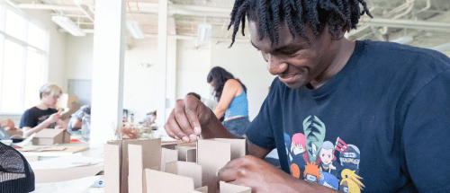Student assembling a cardboard architectural model at a table in a bright Norwich University classroom, with others working in the background.
