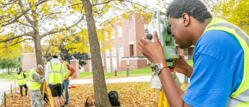 Norwich University students in safety vests conduct a land surveying exercise with a total station, a modern surveying instrument, on a leafy fall campus lawn near a brick academic building.
