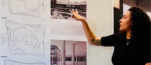 Person with curly hair and a cross necklace presents architectural plans and building photographs on a wall, gesturing with an arm that reveals a leafy tattoo.