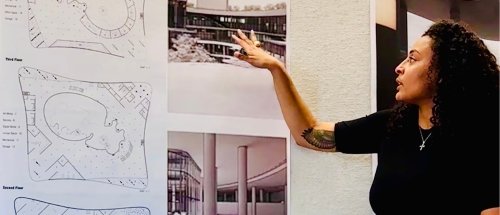 Person with curly hair and a cross necklace presents architectural plans and building photographs on a wall, gesturing with an arm that reveals a leafy tattoo.