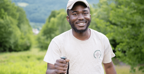 A man smiles at the camera while hiking on a green forested trail, holding a trekking pole and wearing a light-colored T-shirt and cap.