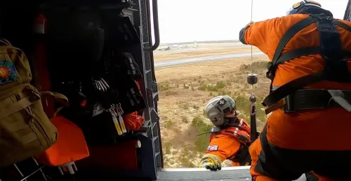 Rescue crew members in bright orange suits and helmets preparing for operation from open helicopter door, looking down at rural landscape below.