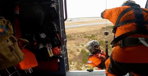 Rescue crew members in bright orange suits and helmets preparing for operation from open helicopter door, looking down at rural landscape below.