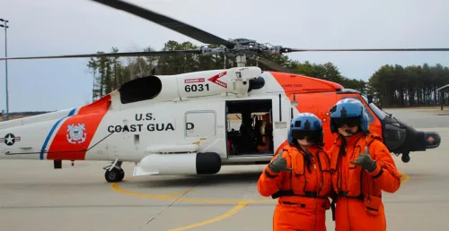 Two people in orange flight suits and helmets pose in front of a U.S. Coast Guard rescue helicopter on a tarmac, with the aircraft door open and crew visible inside.