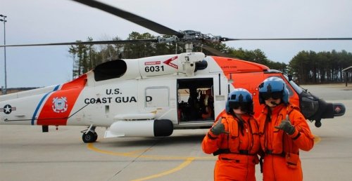 Two people in orange flight suits and helmets pose in front of a U.S. Coast Guard rescue helicopter on a tarmac, with the aircraft door open and crew visible inside.