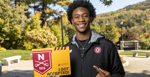 Student holding up accept certificate with Norwich campus behind him.