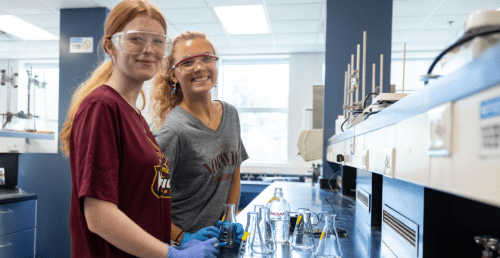 Two students with protective glasses smiling while standing in chemistry class working with science beakers and glasses.