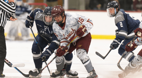Hockey players in digital camo, and navy jerseys battling for puck, intense struggle at face-off circle.