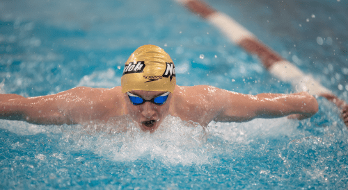 Norwich swimmer wearing a gold cap and blue goggles powers through the water in a butterfly stroke, creating a splash.