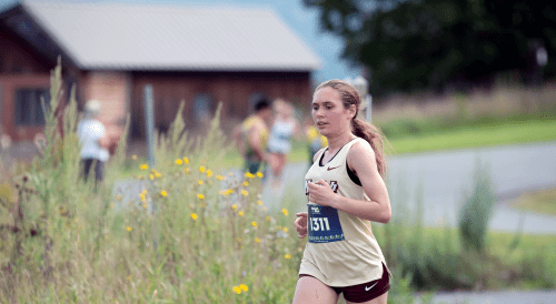 Runner in Norwich jersey, number 1311, racing past yellow wildflowers and a rural building.