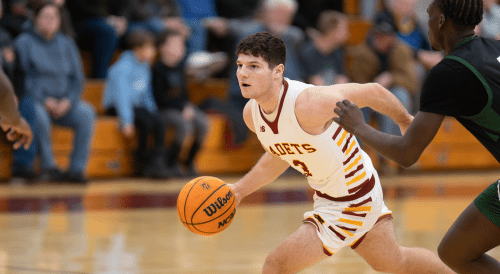 Basketball player in white Cadets jersey dribbling past defender during game with spectators in gymnasium background.