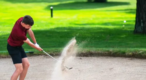 Golfer in maroon shirt hitting out of sand bunker with explosion of sand visible as club strikes through the sand on green golf course.