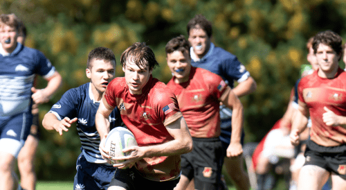 Rugby player in red evades defenders in blue, sprinting with the ball during a sunny outdoor match.