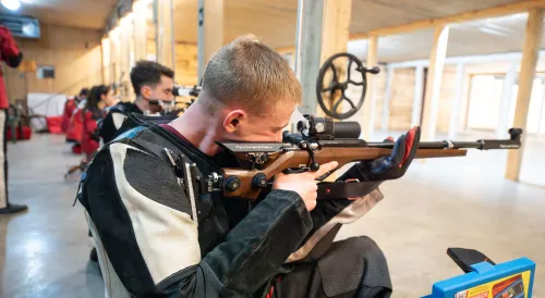 Competitive shooter in specialized jacket aiming a precision air rifle with scope at indoor shooting range, other shooters visible in background.