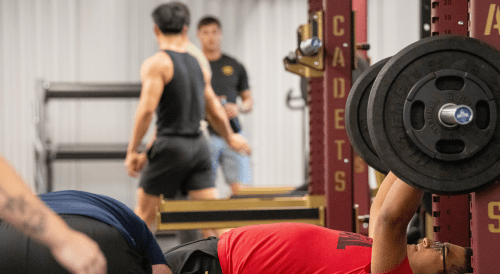Weightlifting session in a gym with barbell rack and weight plates; people training in a cadets fitness facility.
