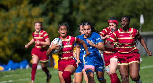 Women's rugby player in red and gold striped jersey carrying ball mid-run, pursued by opponent in blue, with teammates in matching uniforms running behind on sunny field.