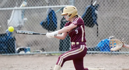 Softball player in maroon uniform with number 3 swinging bat at yellow ball, wearing gold helmet, with catcher's mitt visible and chain-link fence in background.