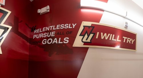 A Norwich University hallway features bold crimson and gold wall graphics. On the left, the NU logo is paired with the motivational phrase “Relentlessly pursue all of your goals” over a faint image of a helicopter. To the right, another NU logo accompanies the school’s motto, “I Will Try,” mounted on a curved sign.