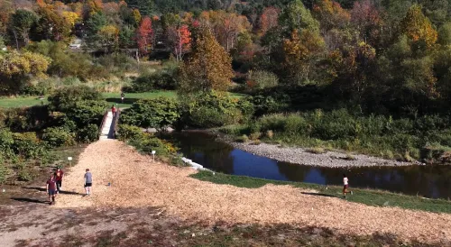 A group of people walk on a woodchip path by a river, surrounded by dense autumn trees; a small footbridge crosses the water in the background.