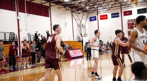 Basketball players in maroon Norwich uniforms and white Trinity uniforms play a game in a gym as teammates and coaches watch from the sidelines.