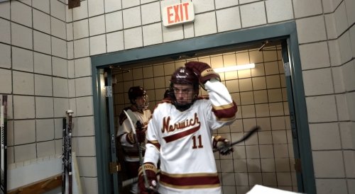 A hockey player in a white and maroon Norwich jersey walks through a doorway under an exit sign.