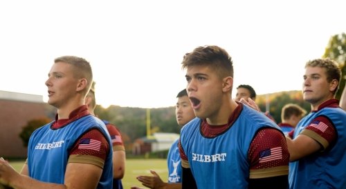 Several young men stand outdoors in blue pinnies with "GILBERT" and US flag patches, clapping and looking forward as the sun sets behind them.