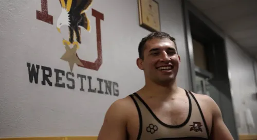 A person wearing a tan and black wrestling singlet stands in front of a wall with a Norwich University wrestling logo.