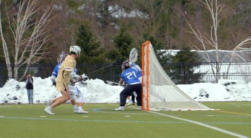 Four lacrosse players, wearing helmets and pads, play near the goal on a green field bordered by snow banks and trees.​