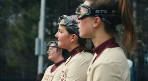 Three female athletes in matching tan and maroon uniforms wear lacrosse goggles.