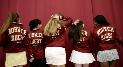 Five women facing away from the camera wear maroon jackets that say "Norwich Rugby," standing in front of a maroon background.