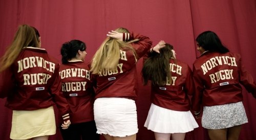Five women facing away from the camera wear maroon jackets that say "Norwich Rugby," standing in front of a maroon background.