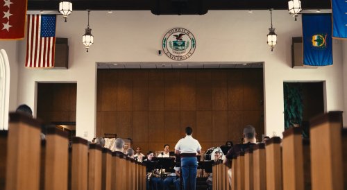 Band conductor leads musicians in a Norwich University chapel as audience sits in wooden pews.