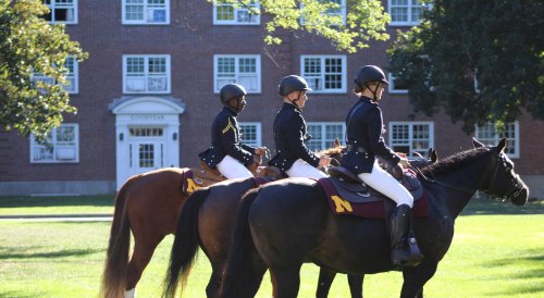 Three riders in uniform sit on horses outside a brick campus building in the afternoon sun.