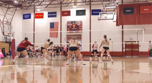 Female volleyball players, wearing tan and maroon uniforms, are engaged in a match in a brightly lit gymnasium decorated with flags and an American flag.