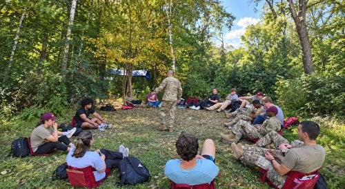 Norwich students gather in a wooded clearing for an outdoor lesson led by an instructor.