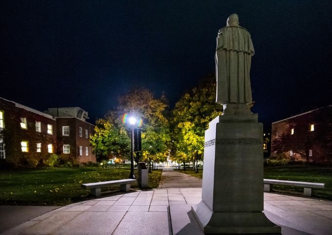 Statue of Alden Partridge at night, illuminated by a streetlamp, with campus pathways, benches, and autumn trees in the background.