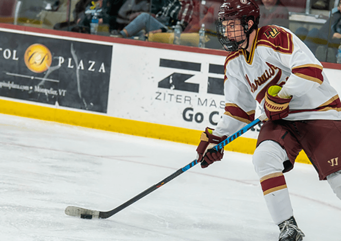 Hockey player in maroon and gold Norwich University uniform skating with puck on ice during a game.