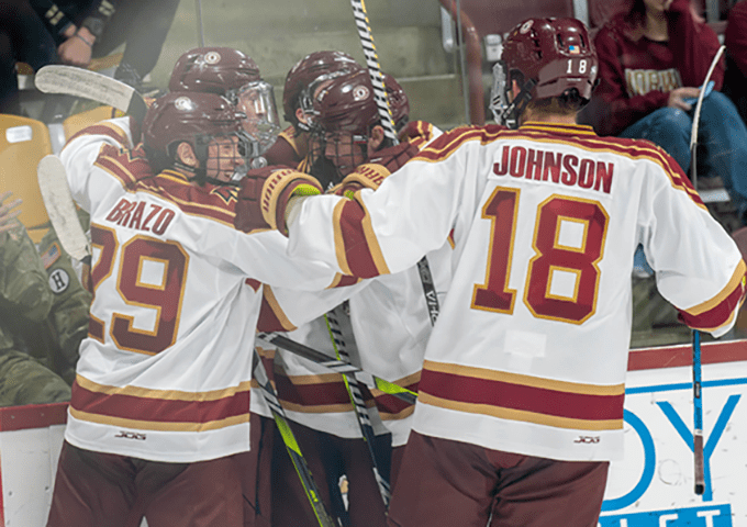Norwich University hockey players celebrate together, with jerseys #29 Crazo and #18 Johnson visible from behind as they huddle with teammates.