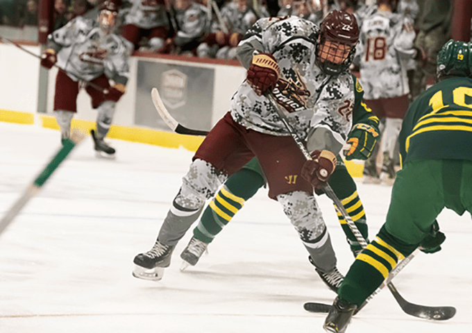 A hockey player in digital camouflage and maroon uniform skating with stick extended near the goal net during a game.