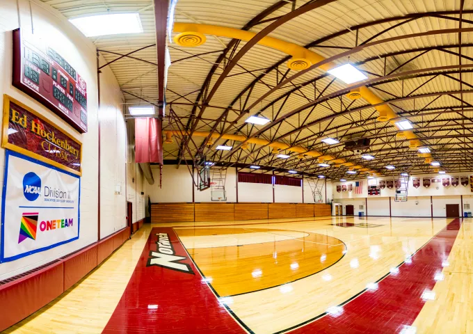 A wide-angle view of an empty Norwich basketball gym, featuring a polished wooden court, red accents, banners, and NCAA Division III signage.