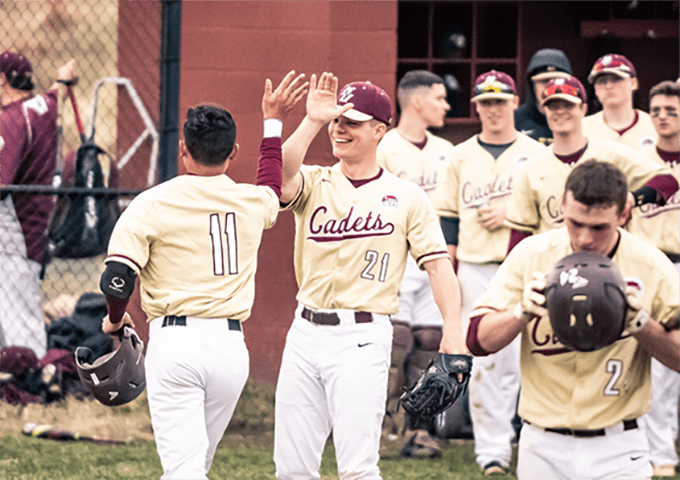 Two baseball players in cream and maroon Cadets uniforms high-five near the dugout while teammates watch and smile in the background.
