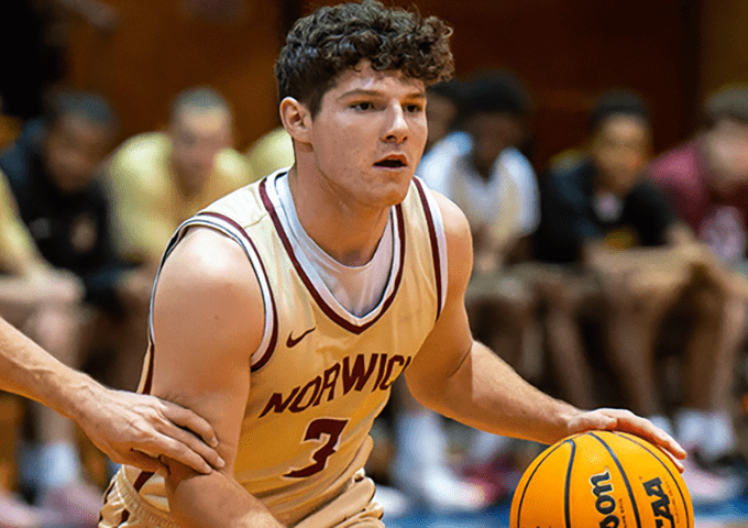 A player in a maroon and gold uniform with text that reads "Norwich", dribbling the ball while looking ahead, with a blurred crowd in the background.
