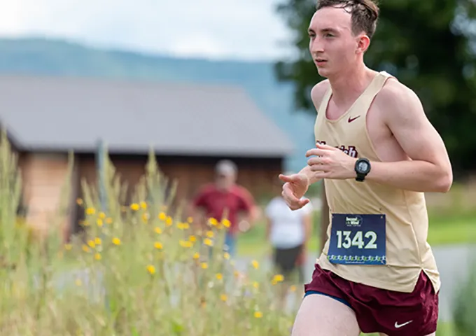 College runner in tan Norwich singlet with race number 1342 competing on rural cross country course with mountains in background.