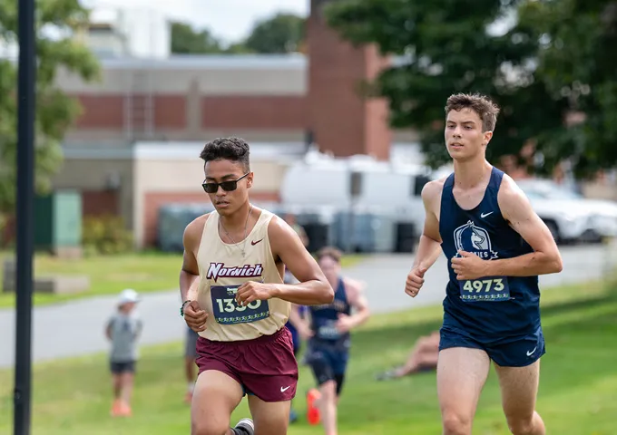 Two college runners competing in cross country race, wearing Norwich and navy uniforms with race numbers on grassy course.