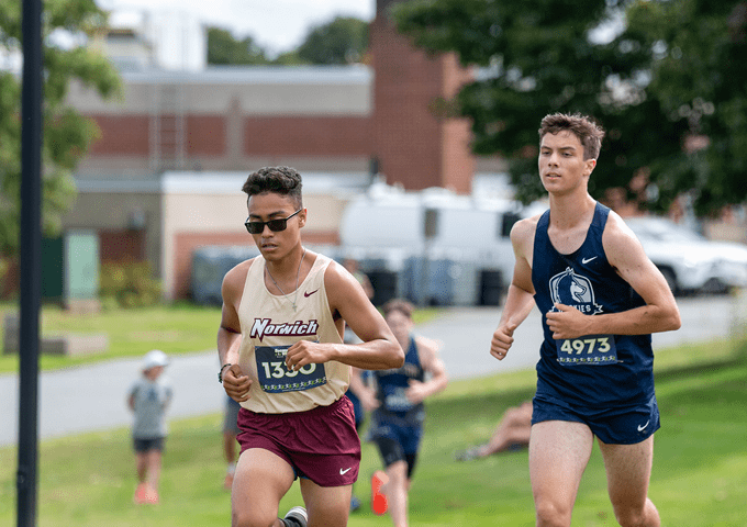 Two college runners competing in cross country race, wearing Norwich and navy uniforms with race numbers on grassy course.