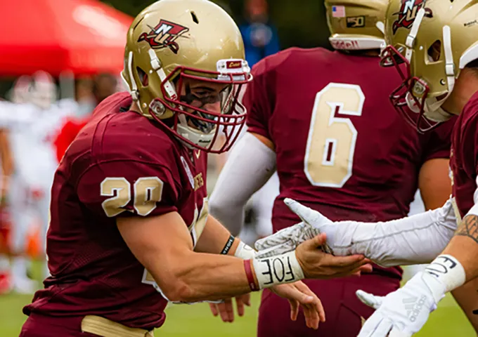 College football players in maroon and gold uniforms celebrating together, with player number 20 in foreground and number 6 visible on teammate's jersey.