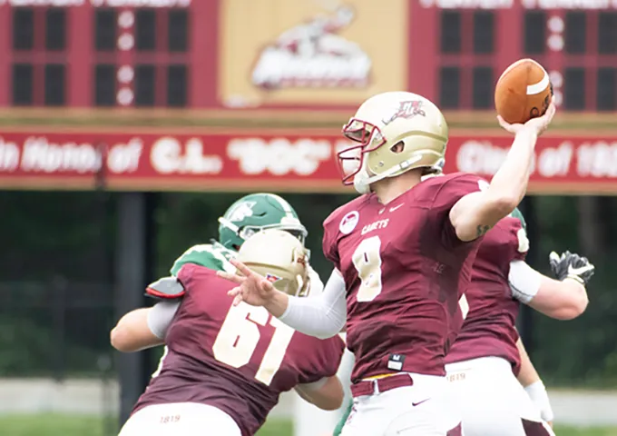 Quarterback in maroon uniform throwing football while teammate number 61 blocks defender in green jersey.