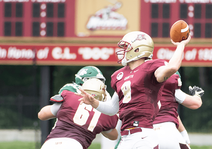 Quarterback in maroon uniform throwing football while teammate number 61 blocks defender in green jersey.