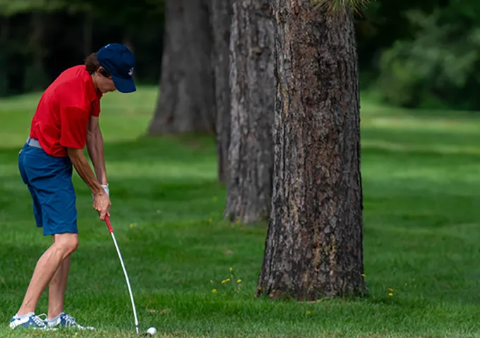 A golfer in a red shirt and blue cap is preparing to hit a ball carefully positioning themselves to navigate around the tree obstacle while on the grass.